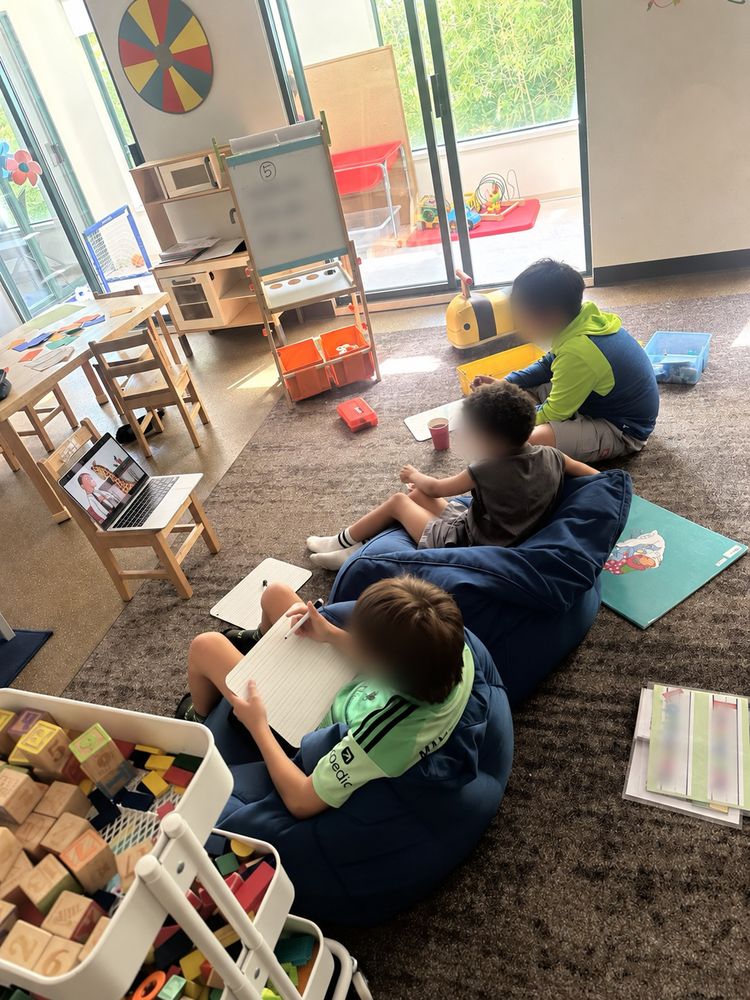 Children seated on beanbags with notebooks, watching a shared screen activity. Faces are blurred.