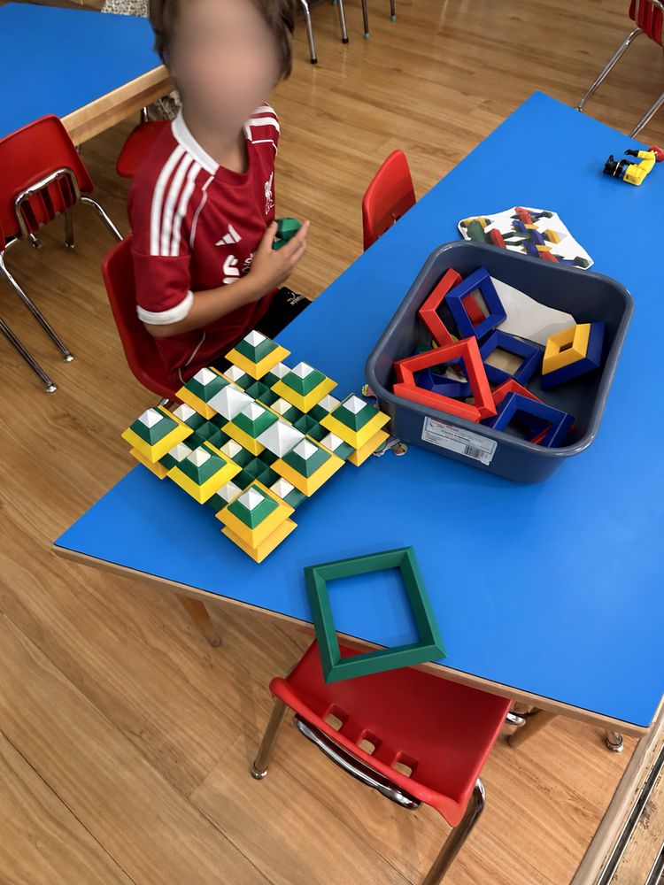 A child building a geometric pattern with coloured shape blocks at a table. Face is blurred.