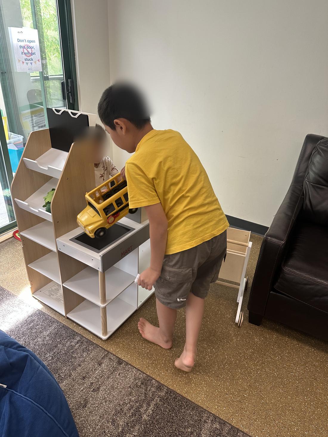 A child playing with a toy bus at a play shelf. Faces are blurred.
